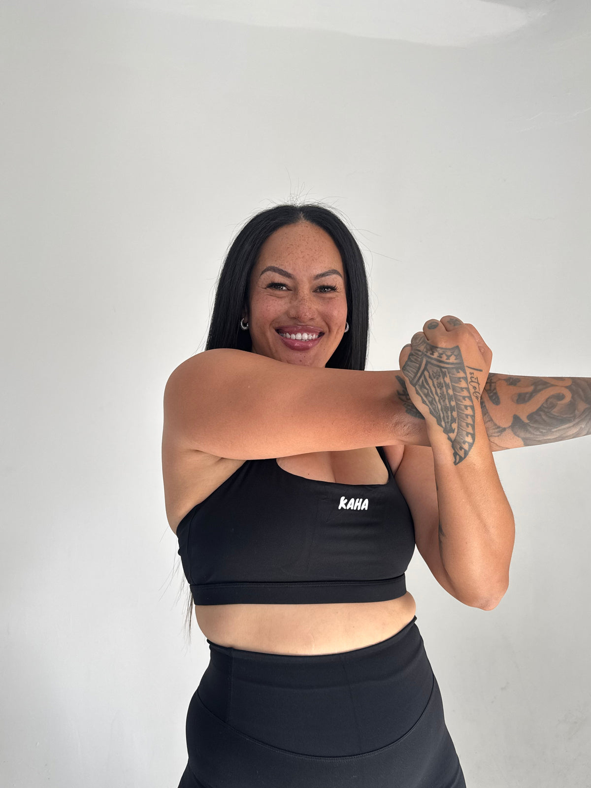 A smiling woman wearing a black KAHA athletic crop top with a square neckline, stretching her arms in a studio.