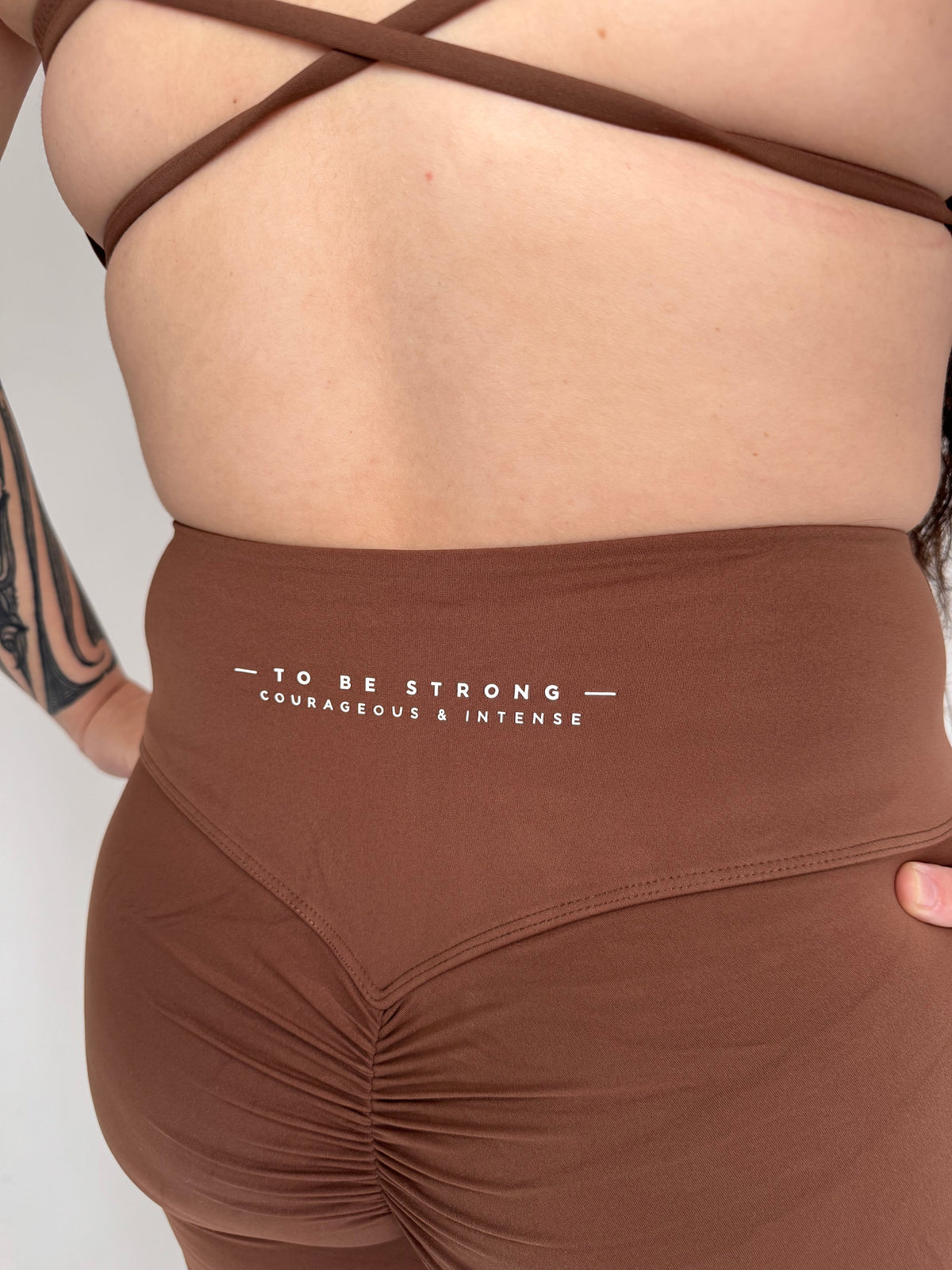 KAHA brown high-waisted flare leggings with back ruching and To Be Strong branding, paired with a criss-cross bra.