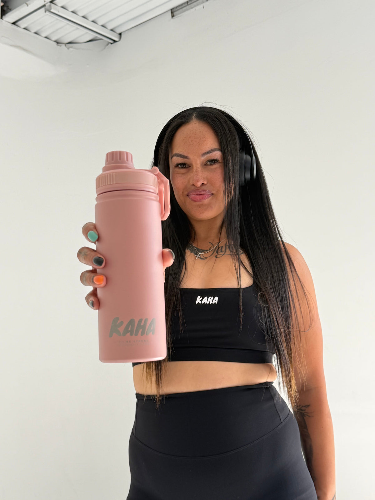 A woman holding a matte pink KAHA insulated stainless steel water bottle with a carry handle in a studio setting.